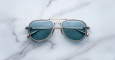 A pair of Jacques Marie Mage Cougar aviator sunglasses with blue lenses and silver frames rests on a white marble surface.