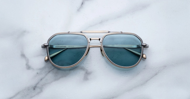 A pair of Jacques Marie Mage Cougar aviator sunglasses with blue lenses and silver frames rests on a white marble surface.