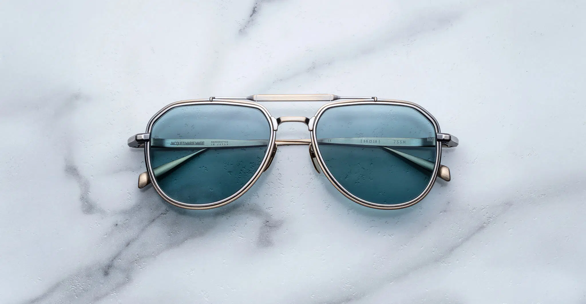 A pair of Jacques Marie Mage Cougar aviator sunglasses with blue lenses and silver frames rests on a white marble surface.