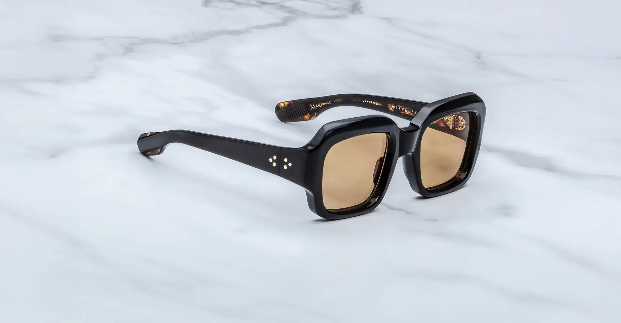 The Jacques Marie Mage Evelyn sunglasses, featuring black square frames and brown-tinted lenses, are displayed on a white marble surface. The arms include subtle gold accents and branding on the inside.