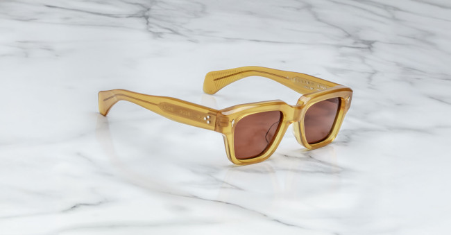A pair of Jacques Marie Mage Fellini square sunglasses with amber frames and brown tinted lenses rests on a white marble surface.