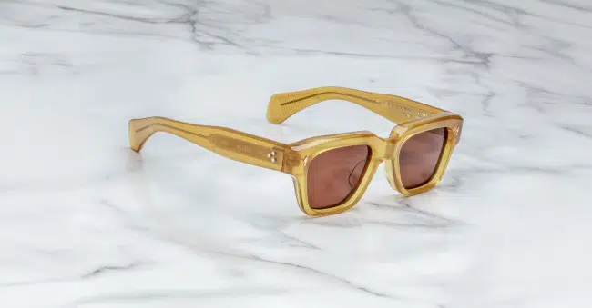 A pair of Jacques Marie Mage Fellini square sunglasses with amber frames and brown tinted lenses rests on a white marble surface.