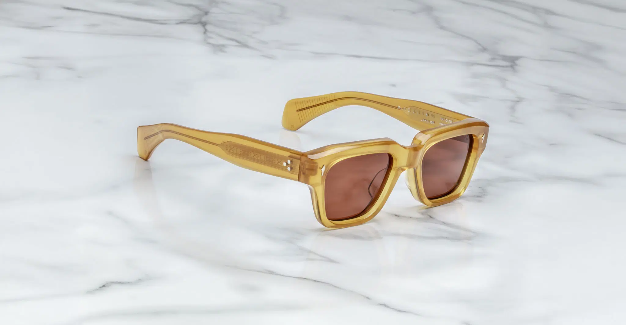 A pair of Jacques Marie Mage Fellini square sunglasses with amber frames and brown tinted lenses rests on a white marble surface.