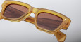 A pair of Jacques Marie Mage Fellini rectangular amber sunglasses with brown tinted lenses is shown in close-up against a light gray background. The arms display engraved branding details.