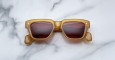 A pair of Jacques Marie Mage Fellini rectangular, amber-colored sunglasses with dark lenses is displayed facing up on a white marble surface with gray veining.