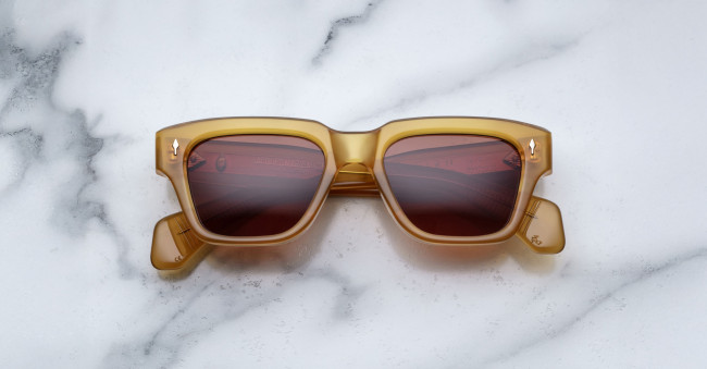 A pair of Jacques Marie Mage Fellini rectangular, amber-colored sunglasses with dark lenses is displayed facing up on a white marble surface with gray veining.