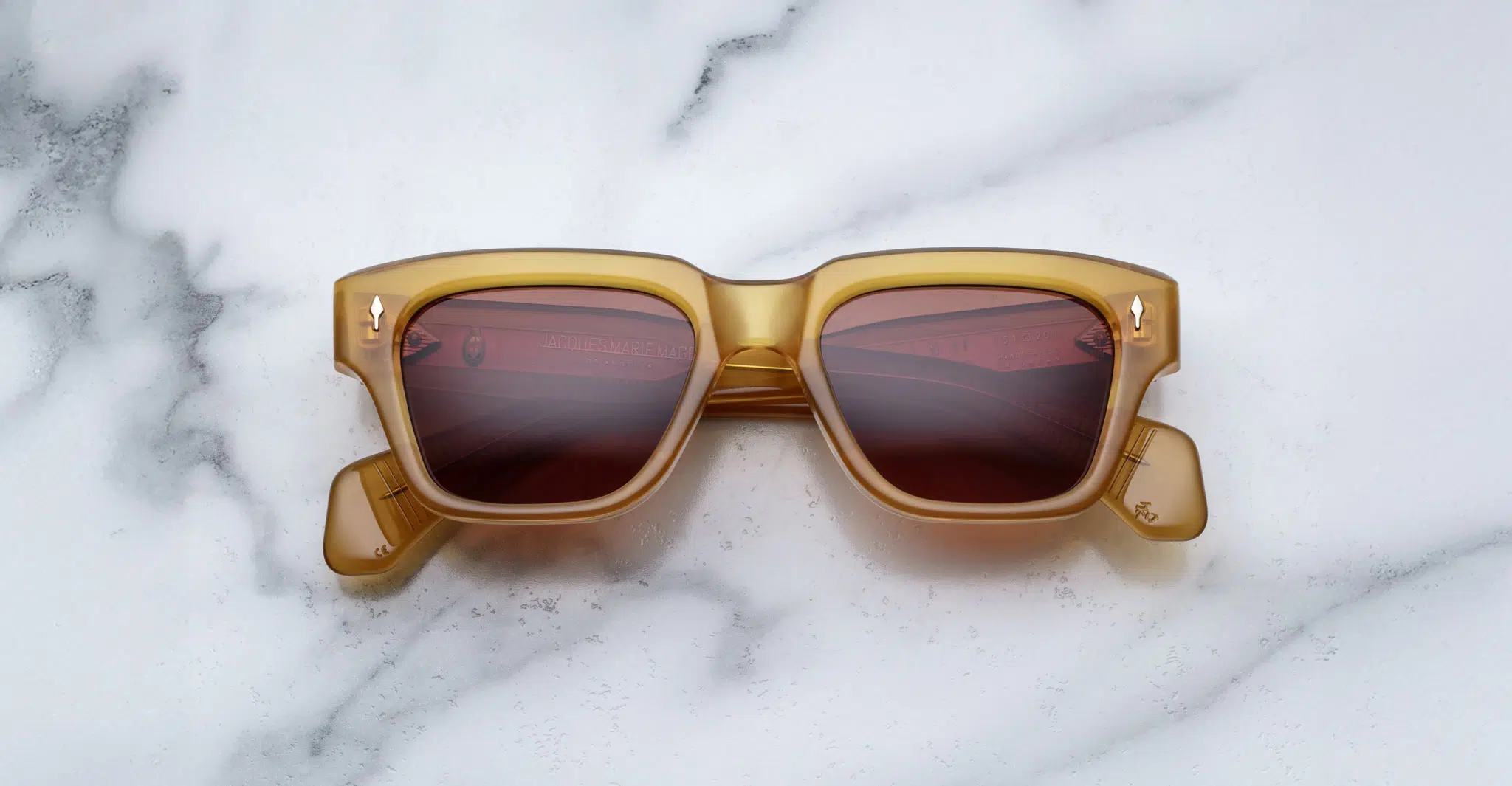 A pair of Jacques Marie Mage Fellini rectangular, amber-colored sunglasses with dark lenses is displayed facing up on a white marble surface with gray veining.