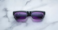 A pair of Jacques Marie Mage Rush black rectangular sunglasses with purple-tinted lenses rests on a white marble surface. Reflections in the lenses hint at partial text and shapes, suggesting the photo was taken indoors.