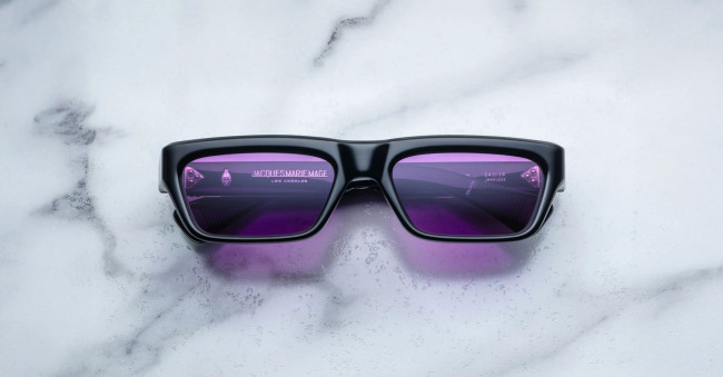 A pair of Jacques Marie Mage Rush black rectangular sunglasses with purple-tinted lenses rests on a white marble surface. Reflections in the lenses hint at partial text and shapes, suggesting the photo was taken indoors.