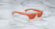 The Jacques Marie Mage Rush sunglasses, featuring an orange rectangular frame with tinted lenses, rest on a white marble surface. Metallic details and decorative elements accent the arms near the hinges.
