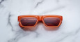 Jacques Marie Mage Rush rectangular sunglasses with thick orange frames and red-tinted lenses rest on a white marble surface. JACQUES MARIE MAGE and LOS ANGELES are visible on the inside of the left lens.