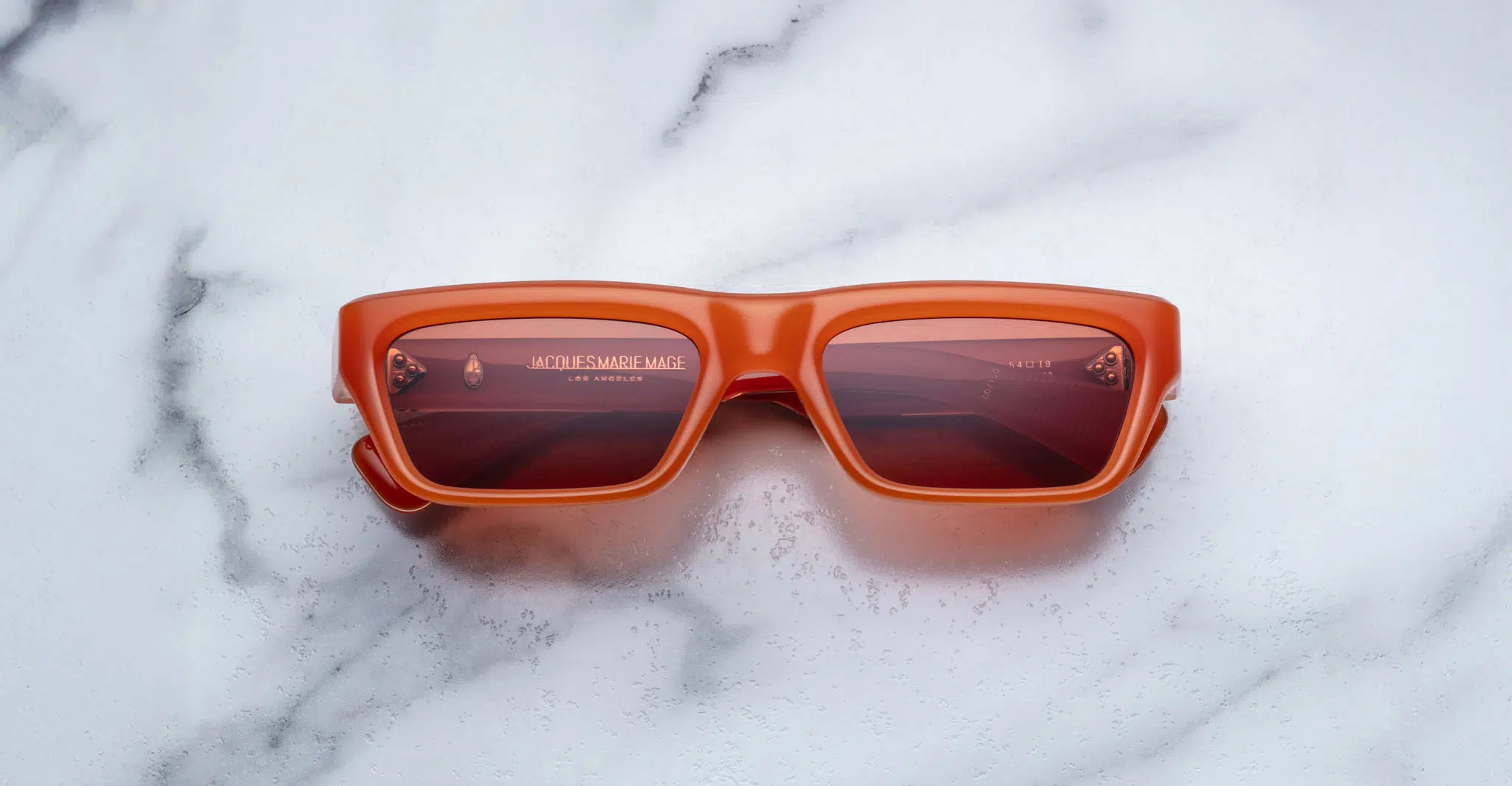 Jacques Marie Mage Rush rectangular sunglasses with thick orange frames and red-tinted lenses rest on a white marble surface. JACQUES MARIE MAGE and LOS ANGELES are visible on the inside of the left lens.
