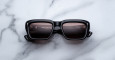 Jacques Marie Mage Walker black square-frame sunglasses with dark lenses rest on a white marble surface, displaying reflections of text and a light source on the lenses.