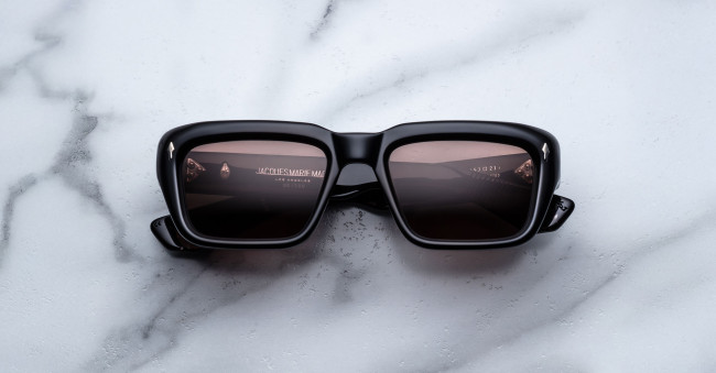 Jacques Marie Mage Walker black square-frame sunglasses with dark lenses rest on a white marble surface, displaying reflections of text and a light source on the lenses.