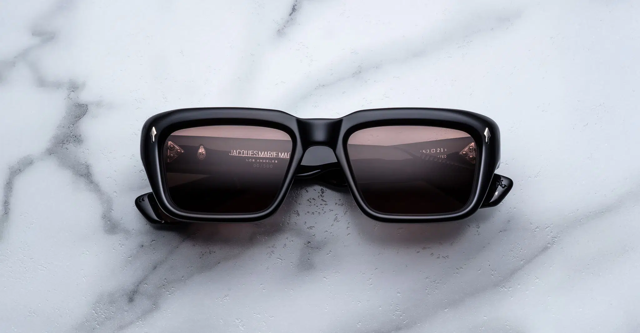 Jacques Marie Mage Walker black square-frame sunglasses with dark lenses rest on a white marble surface, displaying reflections of text and a light source on the lenses.