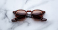 The Jacques Marie Mage Zephirin sunglasses, featuring a brown tortoiseshell square frame and dark lenses, are displayed with folded arms on a white marble surface.