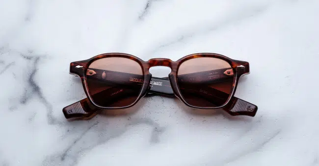 The Jacques Marie Mage Zephirin sunglasses, featuring a brown tortoiseshell square frame and dark lenses, are displayed with folded arms on a white marble surface.