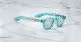 A pair of Jacques Marie Mage Zephirin sunglasses with translucent turquoise round lenses and thick arms rests on a white marble surface.