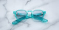 A pair of Jacques Marie Mage Zephirin translucent turquoise eyeglasses with clear lenses is shown folded on a white marble surface.