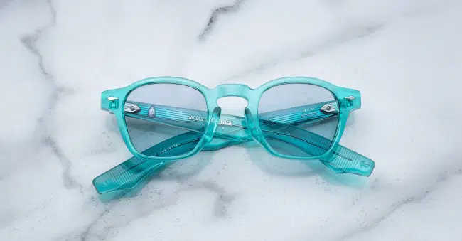 A pair of Jacques Marie Mage Zephirin translucent turquoise eyeglasses with clear lenses is shown folded on a white marble surface.