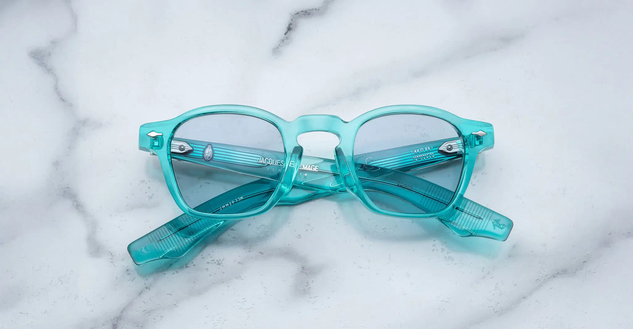 A pair of Jacques Marie Mage Zephirin translucent turquoise eyeglasses with clear lenses is shown folded on a white marble surface.