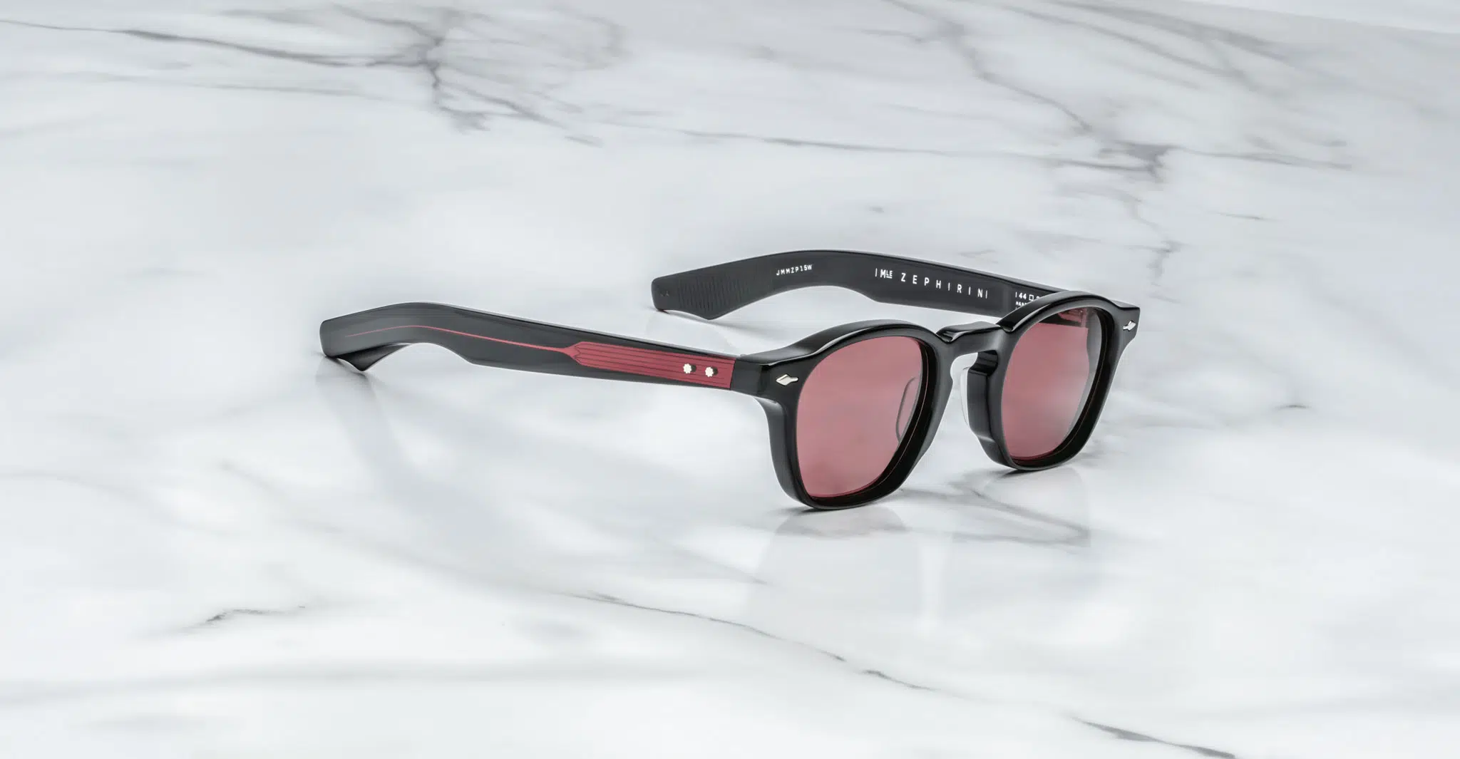 The Jacques Marie Mage Zephirin sunglasses feature black frames, red-tinted lenses, and red detailing on the arms, displayed on a white marble surface.