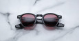 A pair of Jacques Marie Mage Zephirin round, black-framed sunglasses with red-tinted lenses is laid flat on a white marble surface, displaying visible markings and branding on the arms.