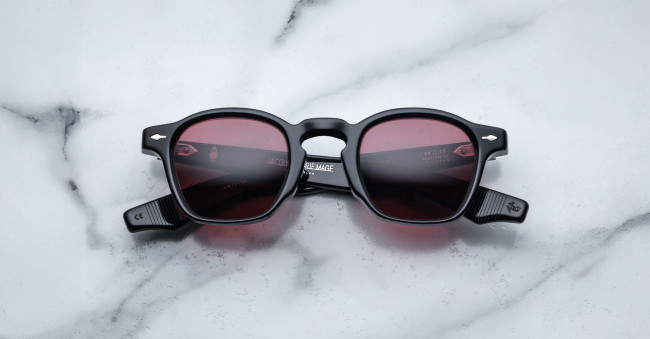 A pair of Jacques Marie Mage Zephirin round, black-framed sunglasses with red-tinted lenses is laid flat on a white marble surface, displaying visible markings and branding on the arms.