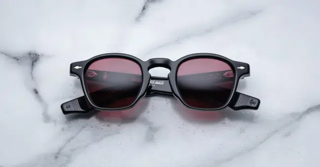 A pair of Jacques Marie Mage Zephirin round, black-framed sunglasses with red-tinted lenses is laid flat on a white marble surface, displaying visible markings and branding on the arms.