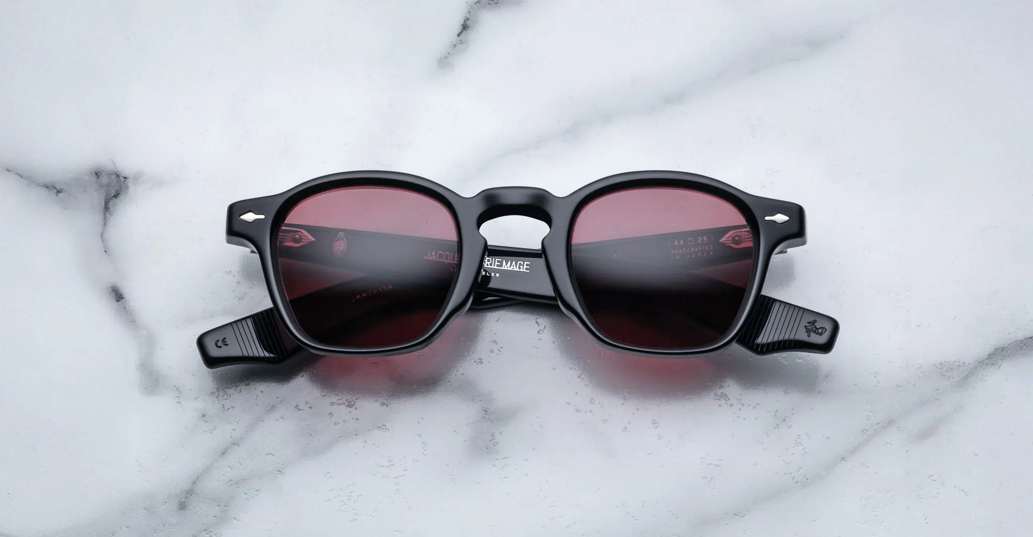 A pair of Jacques Marie Mage Zephirin round, black-framed sunglasses with red-tinted lenses is laid flat on a white marble surface, displaying visible markings and branding on the arms.
