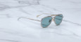 A pair of Jacques Marie Mage 755M aviator-style sunglasses with thin metal frames and blue-tinted lenses rests on a white marble surface.