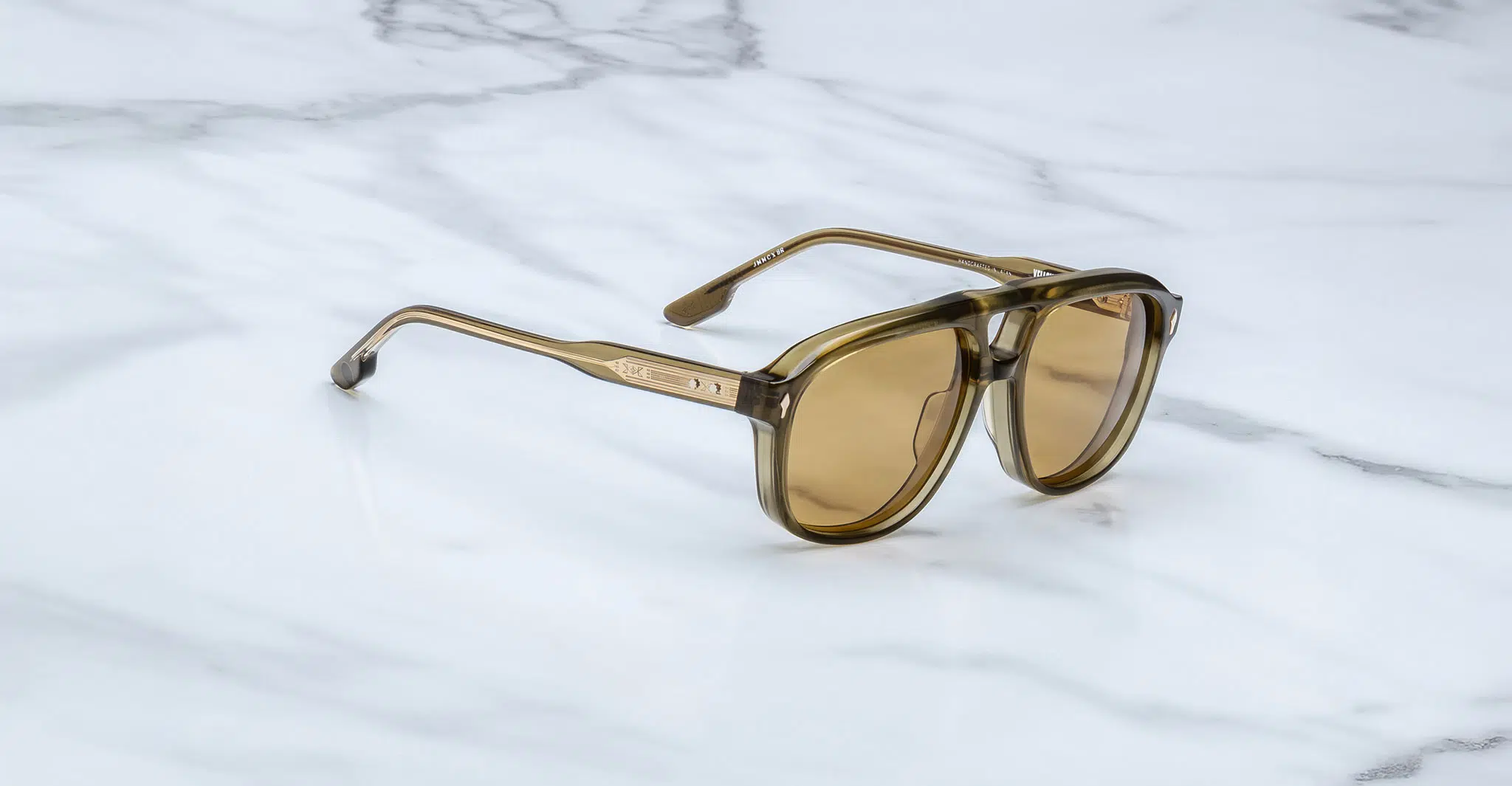 The Jacques Marie Mage Cougar sunglasses, featuring brown-tinted lenses and translucent olive-green plastic frames, are displayed on a white marble surface with grey veining.