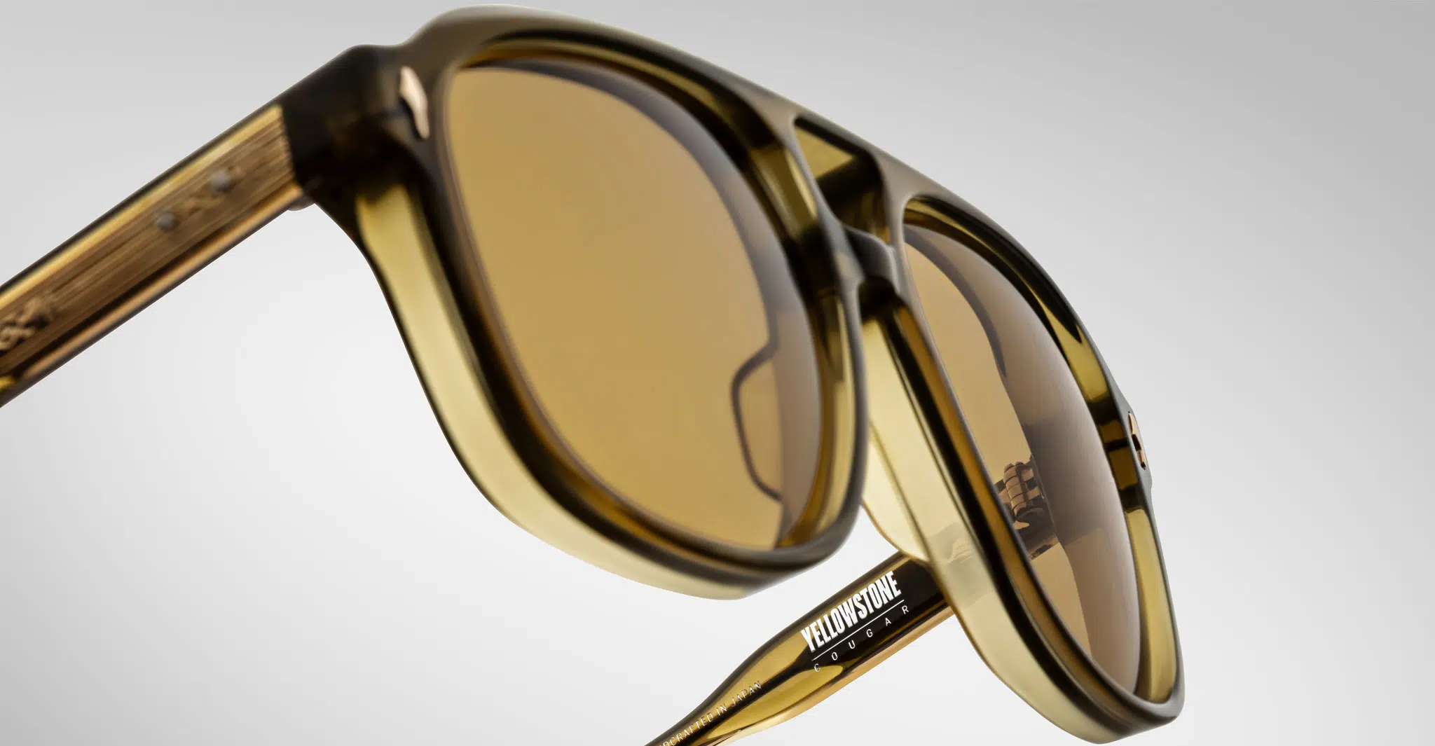 Close-up view of Jacques Marie Mage Cougar translucent brown sunglasses with amber-tinted lenses and a Yellowstone logo on the arm, set against a plain, light-colored background.