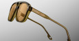 Close-up of Jacques Marie Mage Cougar sunglasses featuring brown-tinted lenses and a thick, transparent olive green frame. The temple arm showcases metallic detailing with two round accents and engraved markings. Set against a plain light gray background.