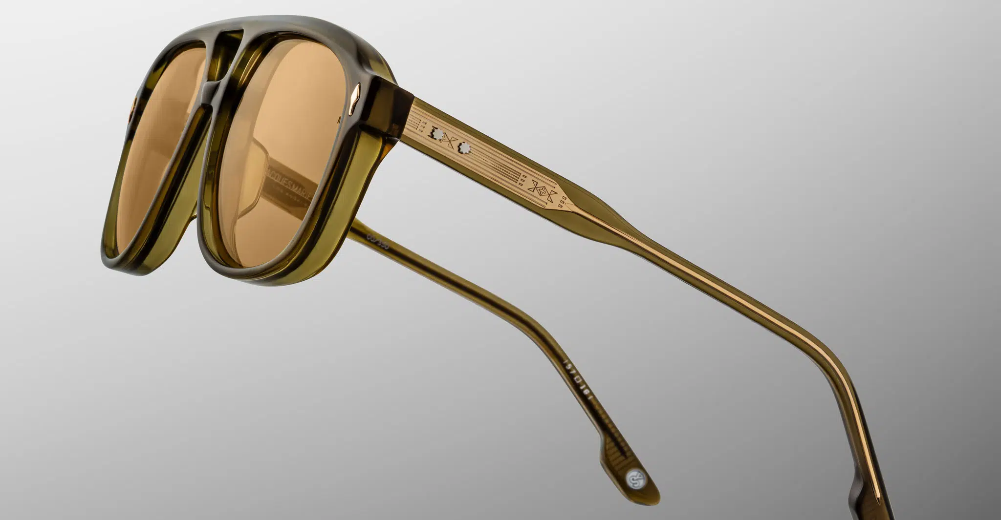Close-up of Jacques Marie Mage Cougar sunglasses featuring brown-tinted lenses and a thick, transparent olive green frame. The temple arm showcases metallic detailing with two round accents and engraved markings. Set against a plain light gray background.