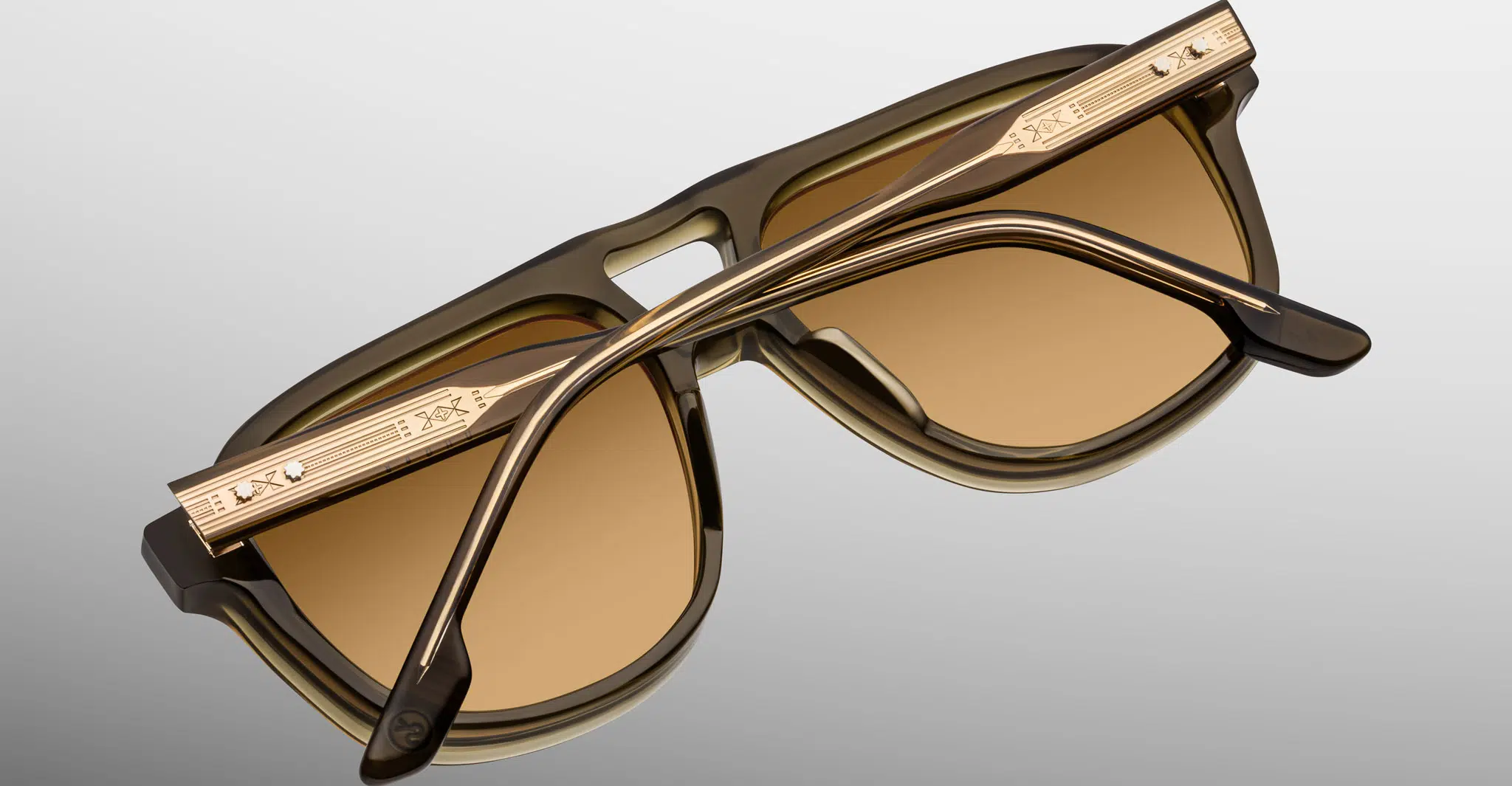 The Jacques Marie Mage Cougar sunglasses feature brown-tinted lenses, gold accents, and decorative temple engravings, displayed with crossed arms over the lenses on a light gradient background.