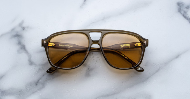 Jacques Marie Mage Cougar olive green sunglasses with amber-tinted lenses rest on white marble with gray veining. Gold text displaying product details appears inside the left temple arm.