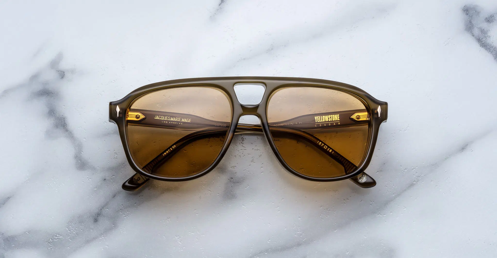 Jacques Marie Mage Cougar olive green sunglasses with amber-tinted lenses rest on white marble with gray veining. Gold text displaying product details appears inside the left temple arm.
