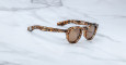 The Jacques Marie Mage Firehole River sunglasses feature round lenses and a brown, black, and beige tortoiseshell frame, displayed on a white marble surface.