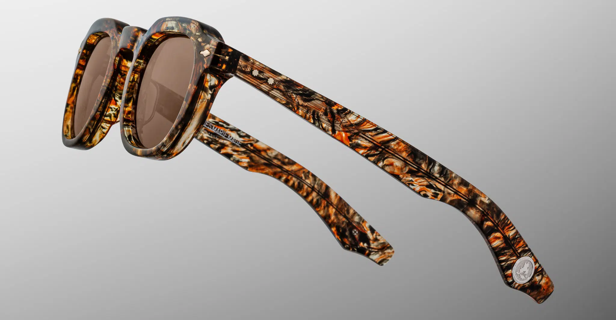 The Jacques Marie Mage Firehole River sunglasses feature brown-tinted lenses and thick, patterned frames in brown, black, and amber, displayed from a side angle against a neutral gray background.