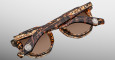 A close-up of two pairs of Jacques Marie Mage Firehole River tortoiseshell sunglasses with brown lenses, stacked diagonally on a light gray background. Both feature metal logo details on the temple hinges.
