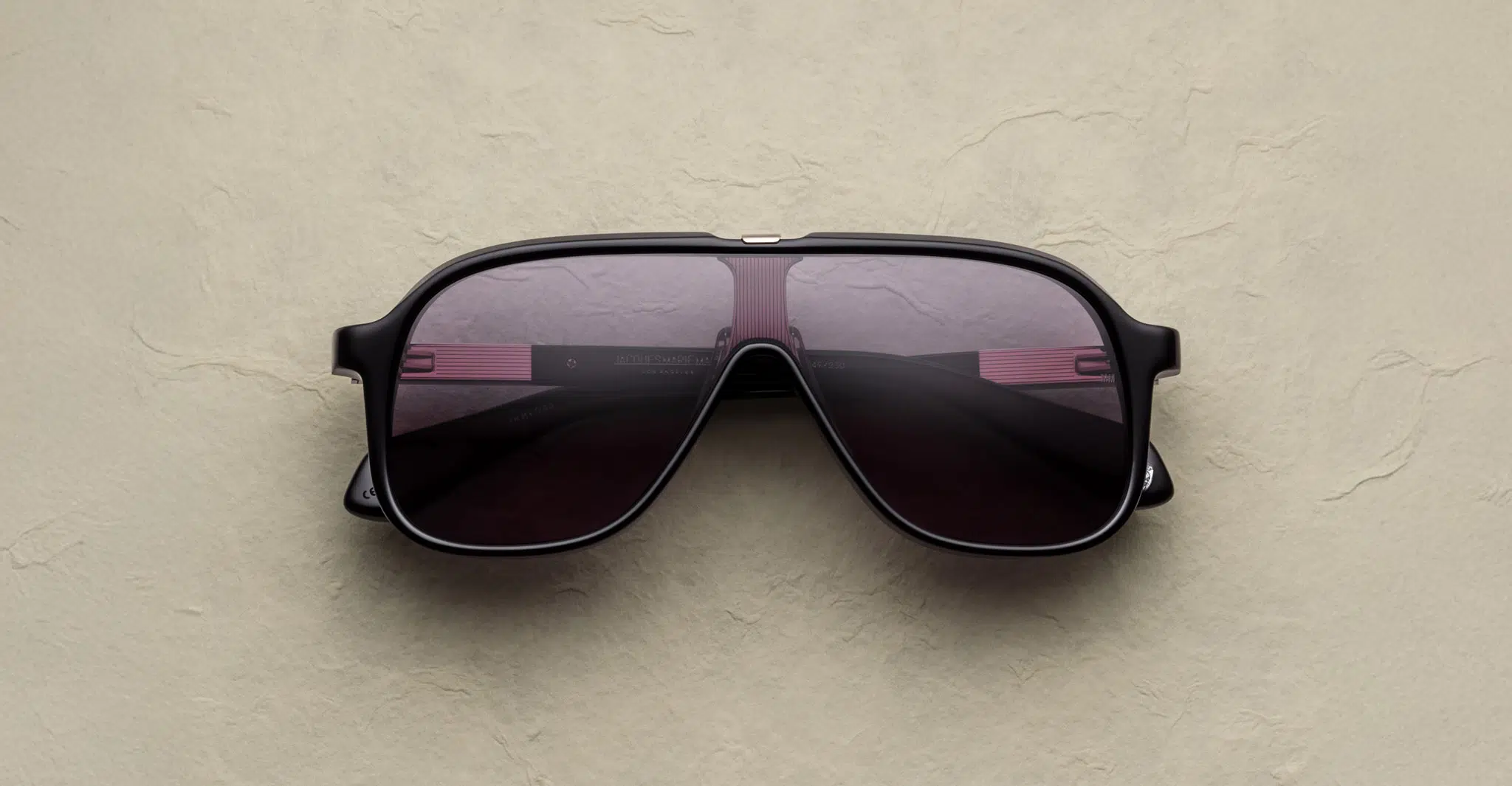 The Jacques Marie Mage Kurokawa sunglasses, featuring a large black aviator design with dark lenses, rest on a beige textured surface.