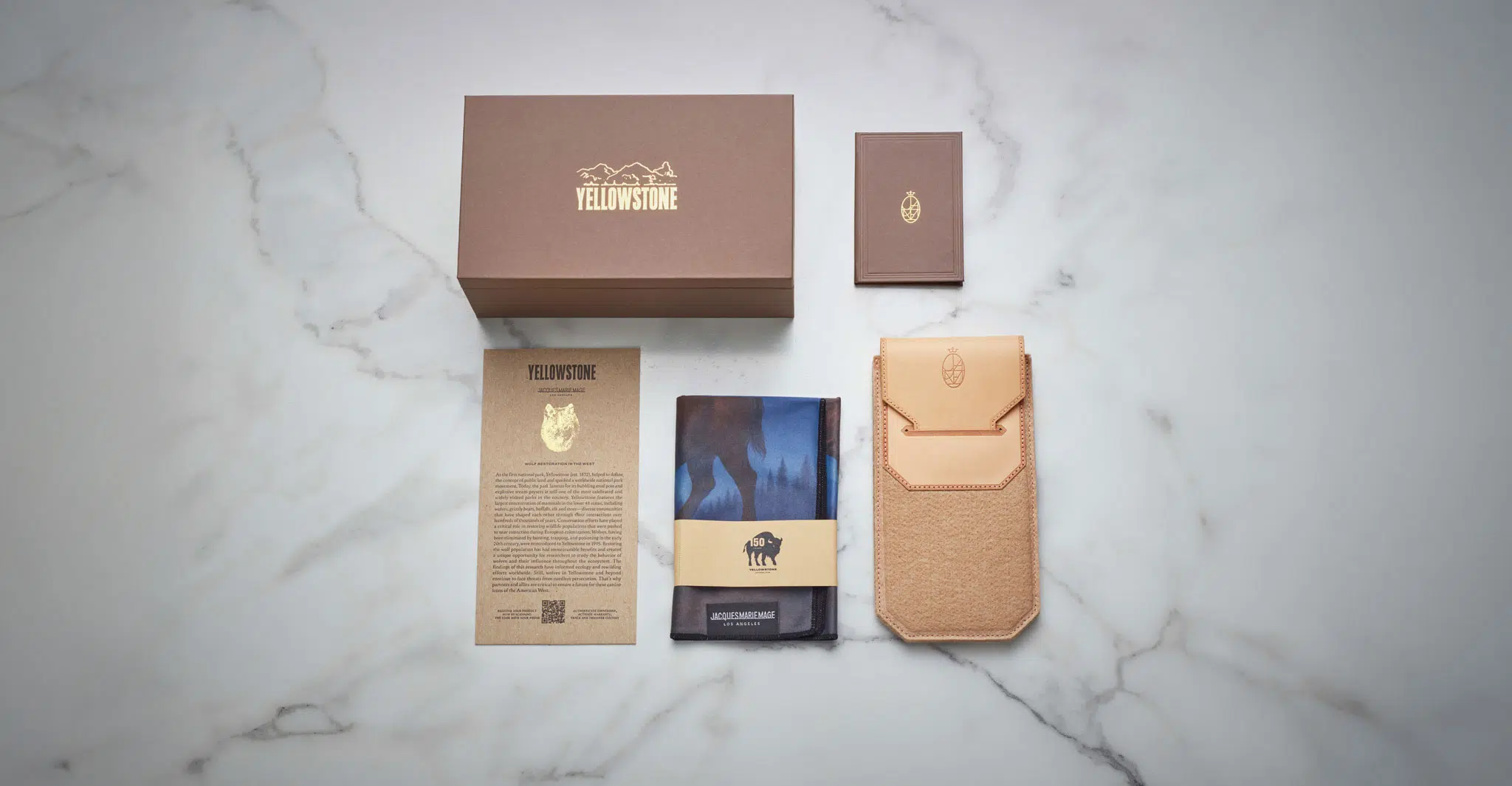 A Jacques Marie Mage Firehole River set—featuring a brown Yellowstone box, small brown booklet, tan leather pouch, card, and folded blue cloth with the Yellowstone bison logo—is displayed on white marble.