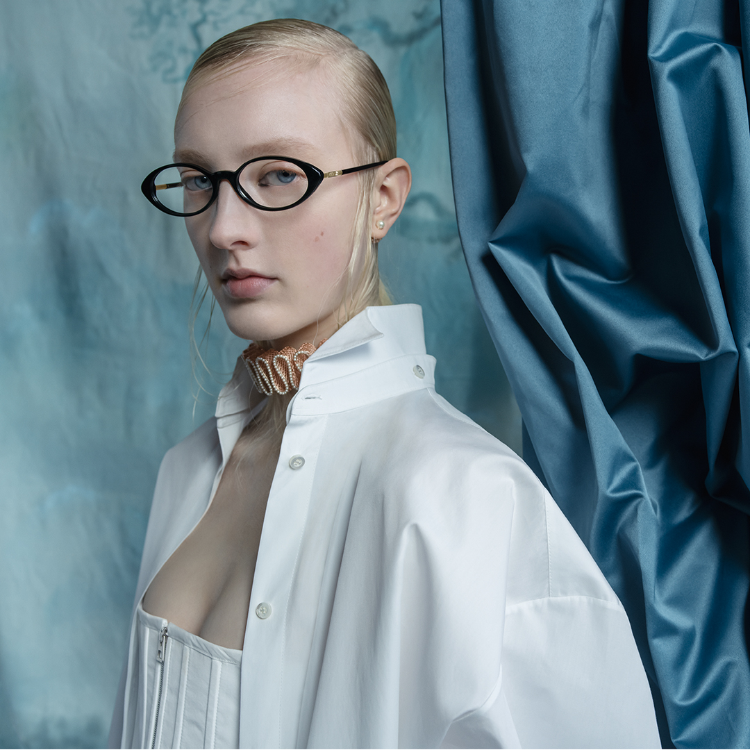 A person with pale skin and blond hair, wearing oval glasses, a white shirt, and a beige choker, stands in front of a blue draped fabric and patterned backdrop, looking slightly to the side.