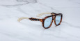 The Jacques Marie Mage Jasper sunglasses feature thick tortoiseshell frames, pale blue lenses, and gold-colored arms, displayed on a white marble surface.