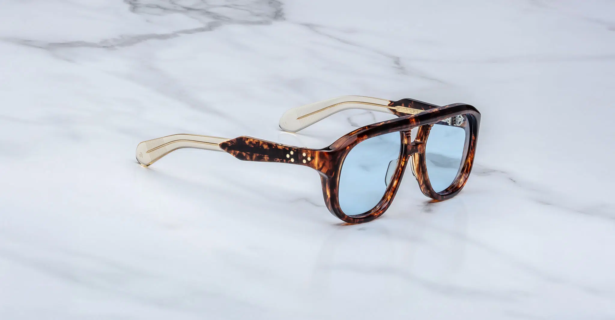 Jacques-Marie-Mage-Jasper-Argyle-JMMYA-5C-3-4-Dan-Deutsch The Jacques Marie Mage Jasper sunglasses feature thick tortoiseshell frames, pale blue lenses, and gold-colored arms, displayed on a white marble surface.