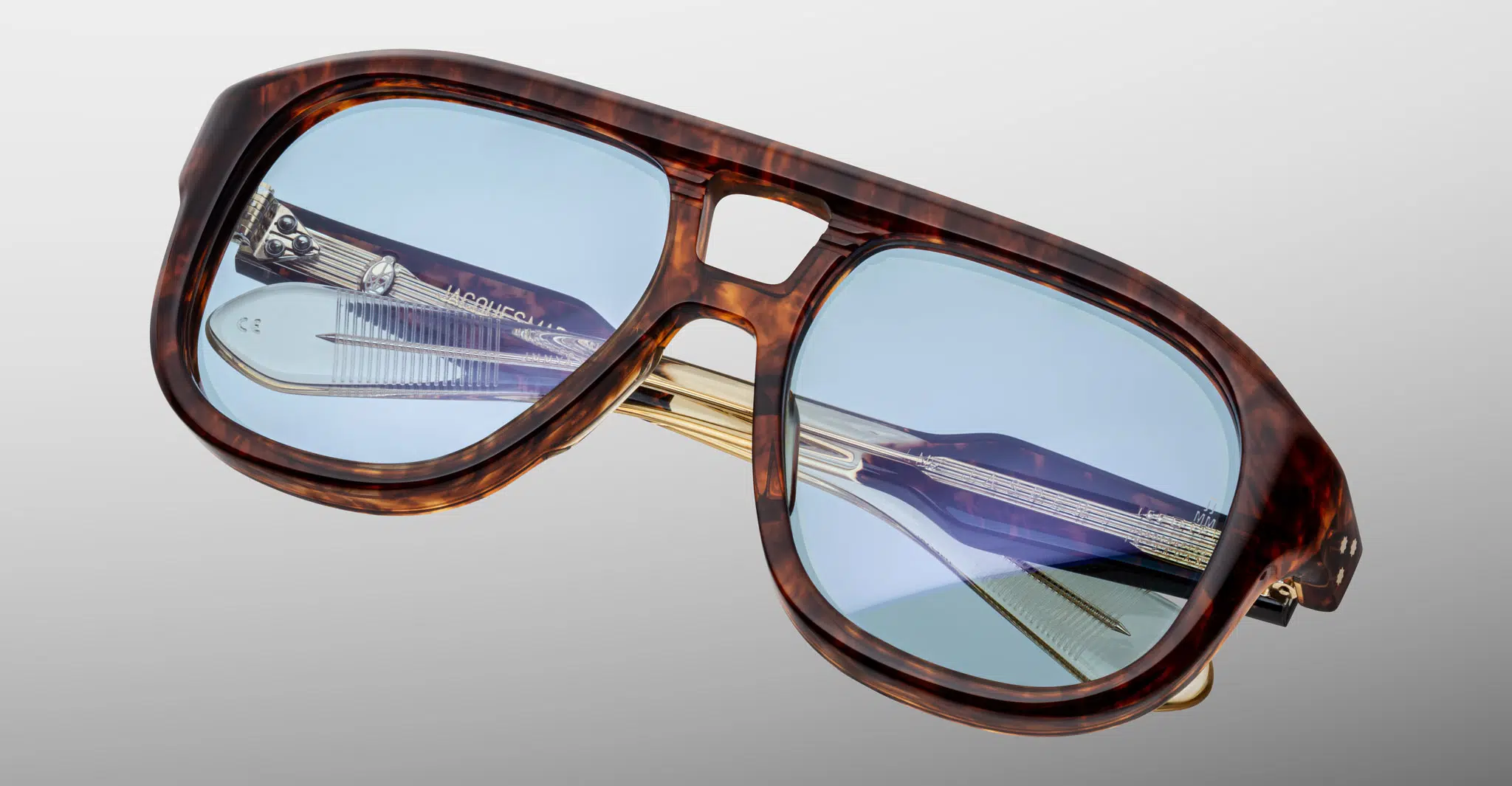 Jacques-Marie-Mage-Jasper-Argyle-JMMYA-5C-Detail1-Dan-Deutsch Close-up of Jacques Marie Mage Jasper oversized sunglasses with a brown tortoiseshell frame, light blue lenses, and metal accents on the nose bridge and temples, set against a neutral gray background.