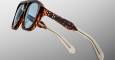 Close-up of Jacques Marie Mage Jasper glasses featuring thick, rectangular blue-tinted lenses, a stylish tortoiseshell frame, and cream-colored arms, shown against a light gradient background.