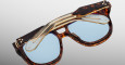 Two closed pairs of Jacques Marie Mage Jasper glasses—one with tortoiseshell frames and blue lenses, the other with gold frames and clear lenses—overlap on a light gray background.