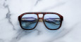 A pair of Jacques Marie Mage Jasper tortoiseshell sunglasses with blue-tinted lenses rests on white marble, reflecting a folded pair of silver-framed eyeglasses in their lenses.